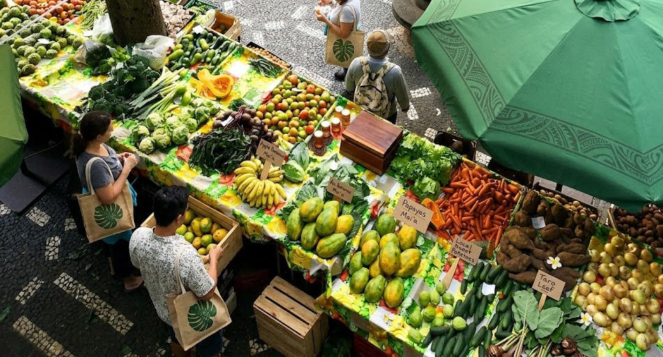 Farmers market stalls heaped with peppers, tomatoes, lettuce, and carrots under a green umbrella